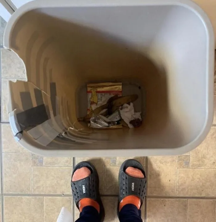 Looking down at feet in slippers in front of a plastic trash can with taped sides, depicting roommate stories.