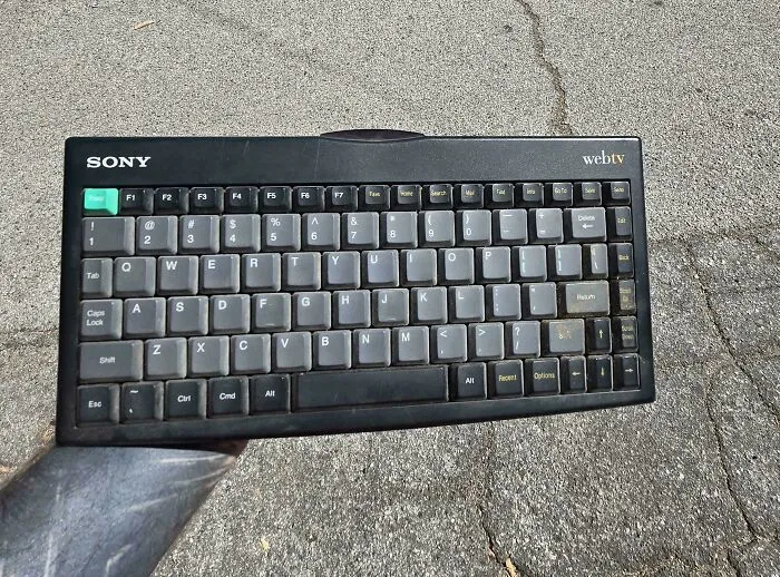 A black Sony WebTV vintage tech keyboard with chunky gray buttons resting on asphalt