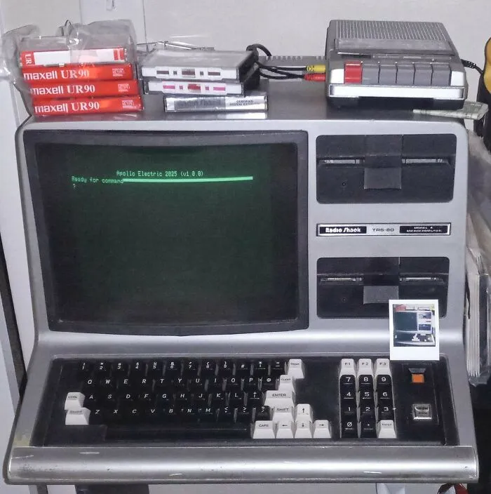 A retro Radio Shack TRS-80 computer surrounded by vintage tech audio cassettes