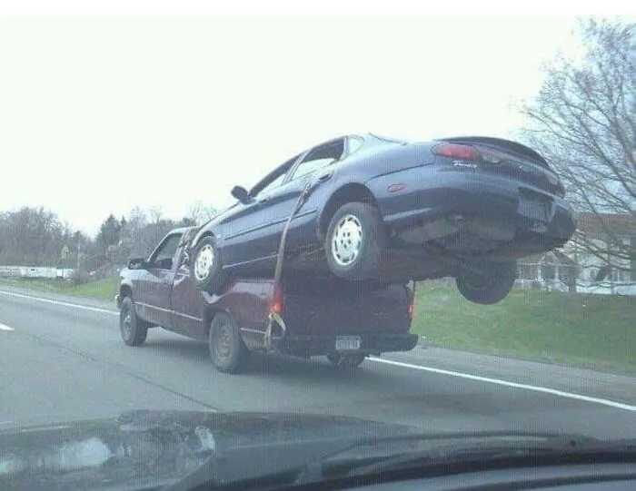 A pickup truck driving on a highway with a car strapped on top of its bed as one of the redneck fixes.
