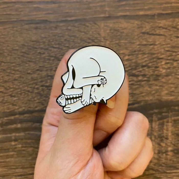 A hand holding a skull enamel pin shaped like a sleeping cat, showing clever ideas from people.