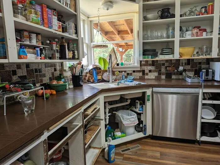 Cluttered home kitchen with exposed under-sink plumbing and open shelving.