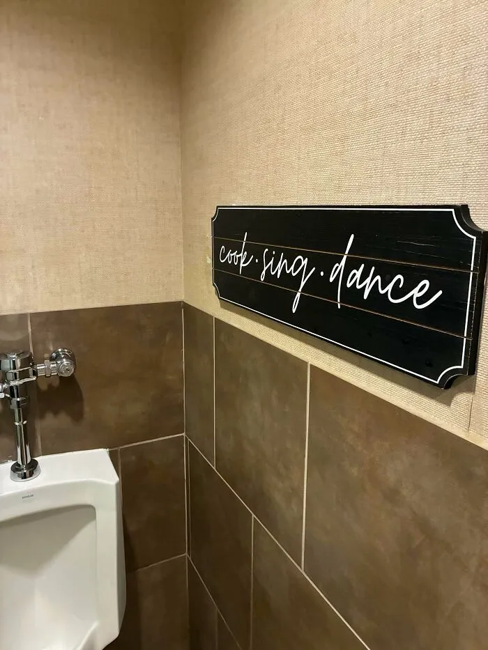 A wooden sign reading cook sing dance placed above a urinal, an odd interior design decision.