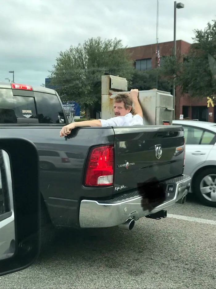 A man sitting in the back of a pickup truck holding a heavy metal object in place, an example of redneck fixes.