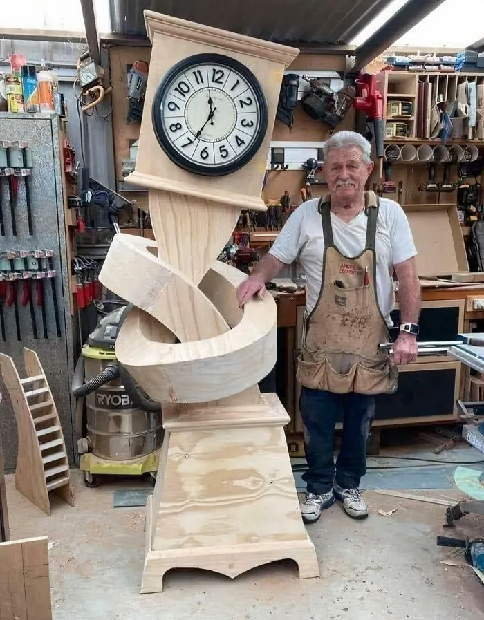 A carpenter stands next to a whimsical grandfather clock with a knotted wooden body, among clever wooden creations