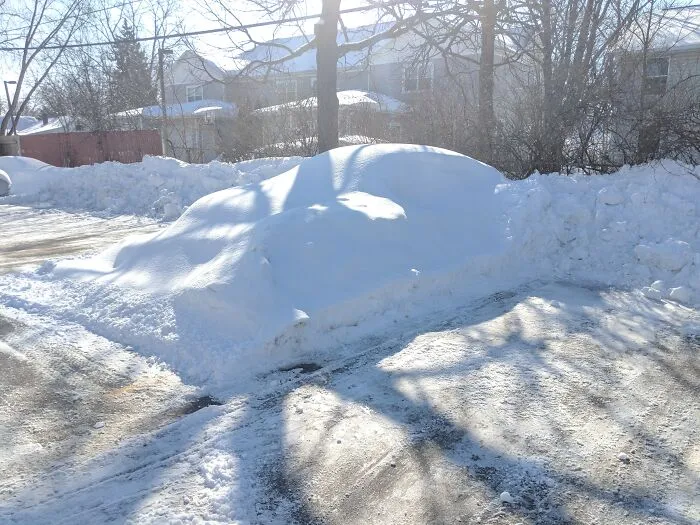 A car completely buried under a thick, smooth layer of white snow in a parking lot, highlighting questionable decisions.