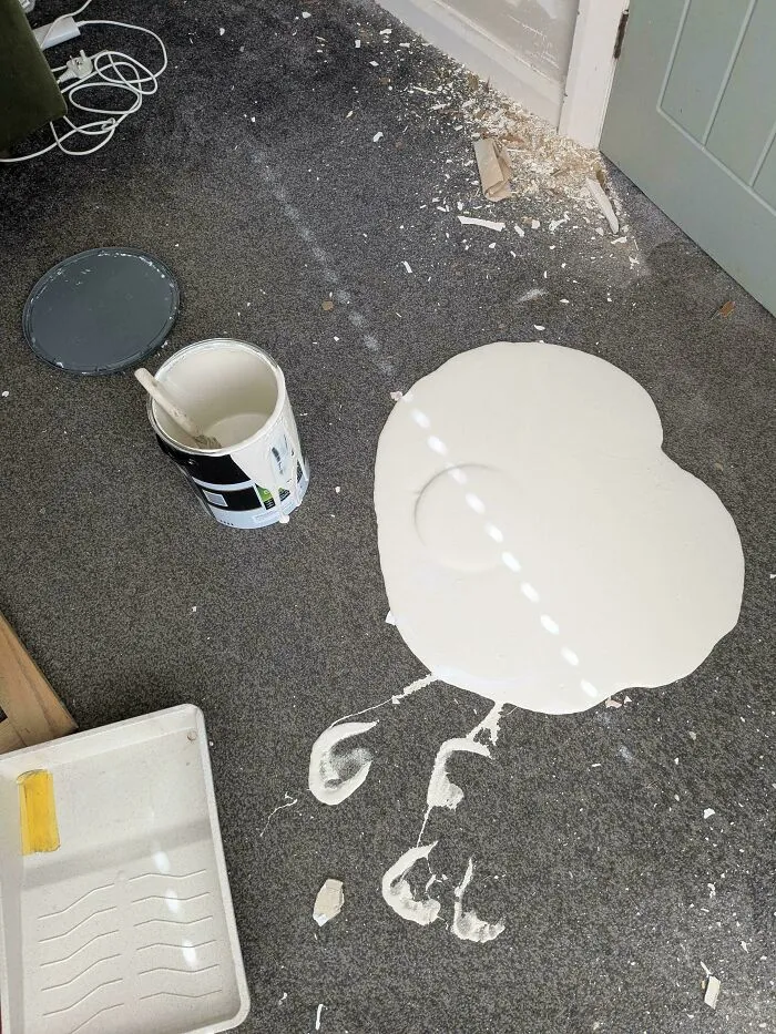 A questionable decision showing white paint spilled on a grey carpeted floor with footprints nearby
