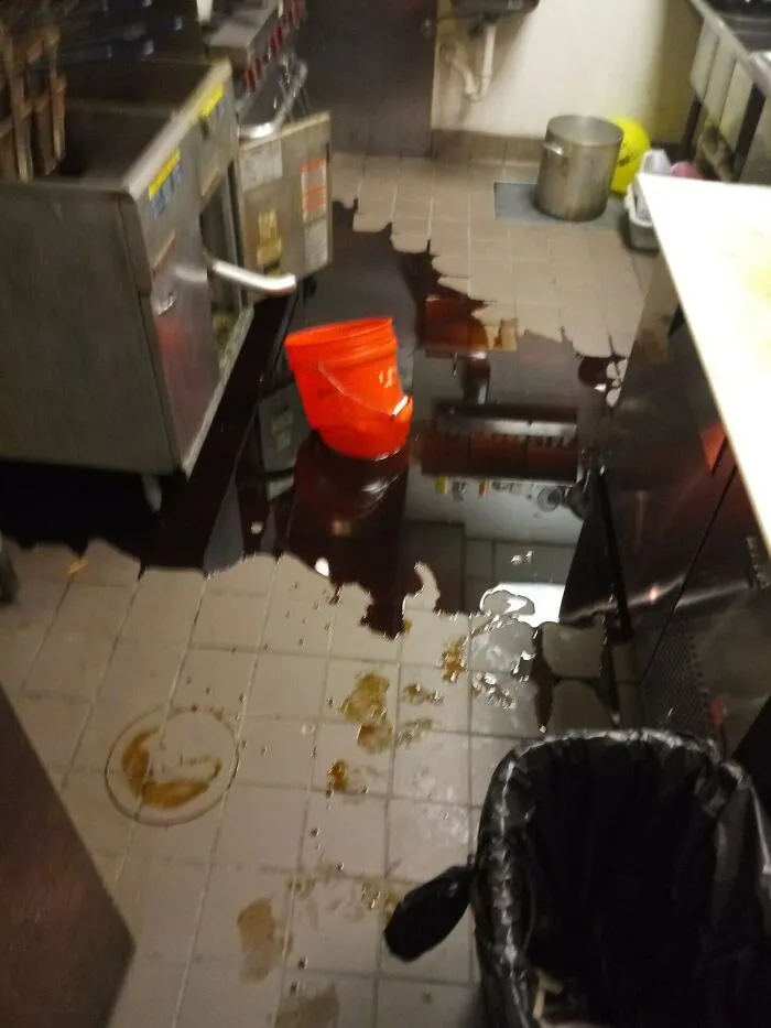 A kitchen floor flooded with dark oil next to deep fryers, showing questionable decisions on maintenance.