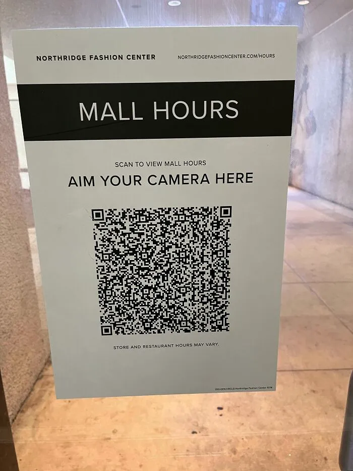 A sign on a glass window saying Aim Your Camera Here with a QR code, an example of questionable decisions.