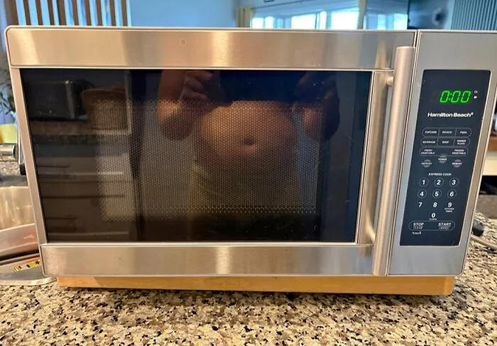 A funny mirror pic reflection of a bare stomach visible on the reflective door of a stainless steel microwave.