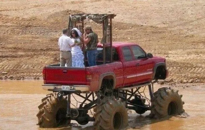 A couple having a questionable wedding ceremony standing in the bed of a monster truck parked in mud.