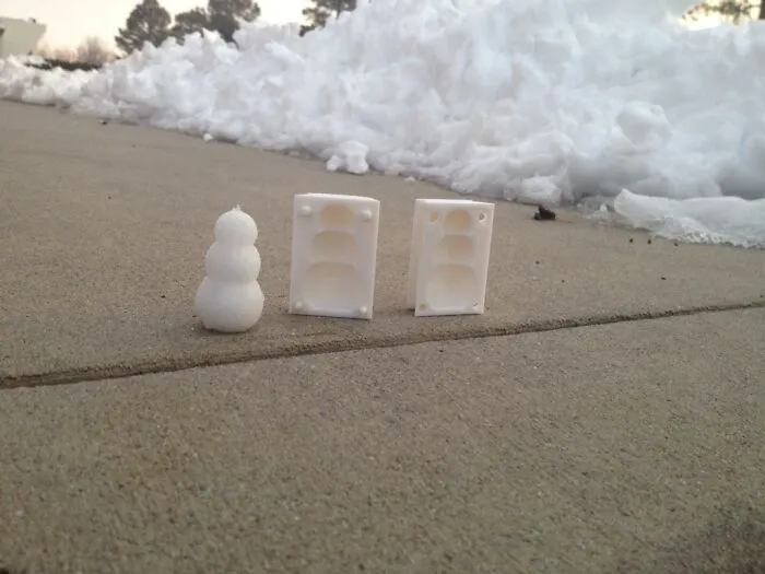 A small snowman next to a two-part 3D printer mold on a concrete sidewalk, showcasing human ingenuity.