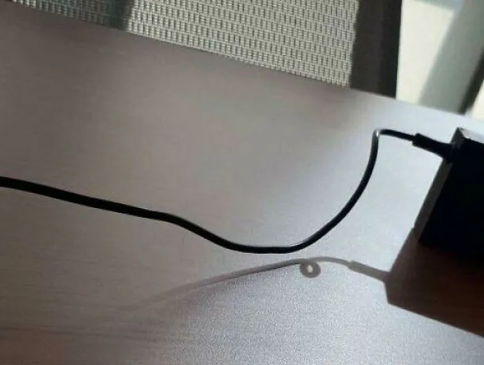 A black power cord on a wooden desk creates a strange photo with a floating loop in its shadow.