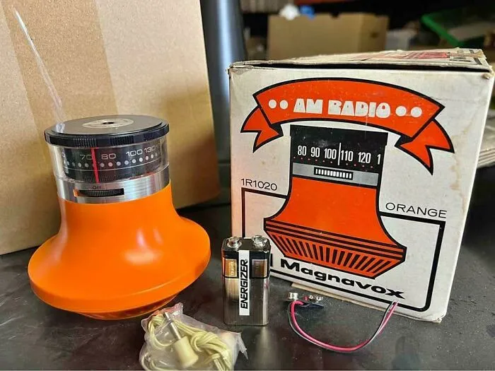 An orange Magnavox AM radio retro gadget standing beside its original product box and a 9V battery.