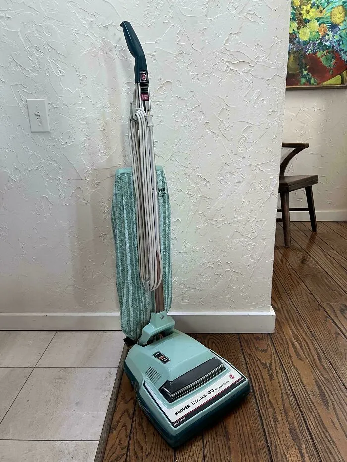 A retro mint green Hoover vacuum cleaner standing against a white textured wall.