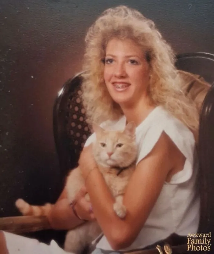 A teenage girl with big hair poses for an awkward family photo holding a calm orange cat
