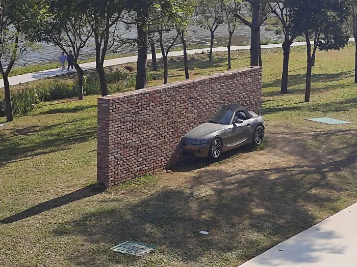 A silver sports car parked right up against a isolated brick wall in a park, showing strange photos.