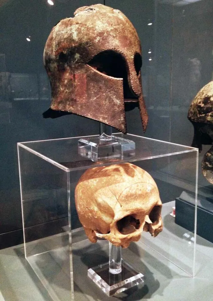 Rusty ancient helmet displayed above old human skull in museum