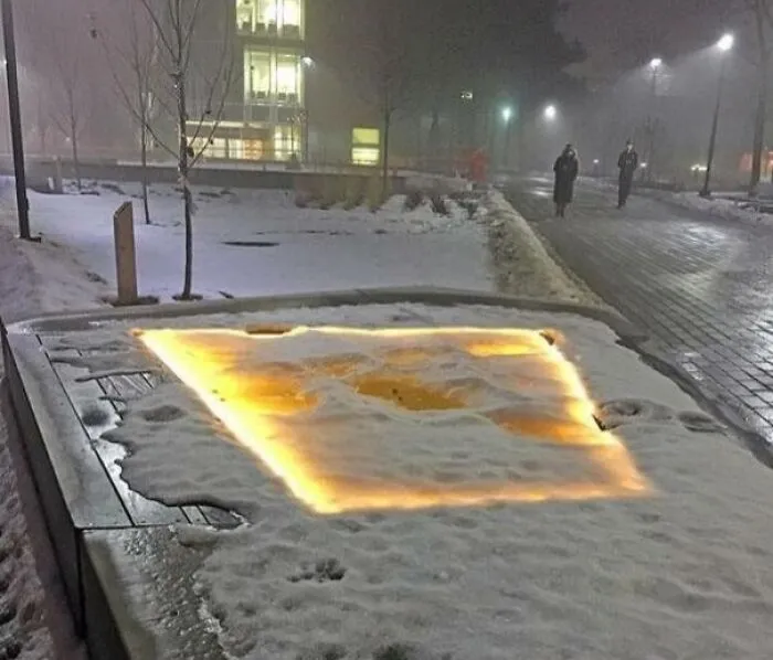 A strange photo showing a glowing yellow light square beneath a layer of snow on an outdoor bench at night.