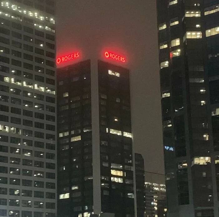 Tall city skyscrapers illuminated at night, featuring a strange photo of two identical glowing Rogers signs.