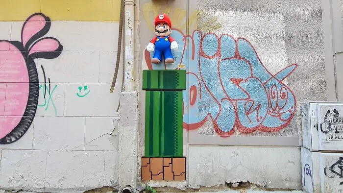 Creative art collective installation showing a 3D Mario figure standing on a green pipe on a city street wall.
