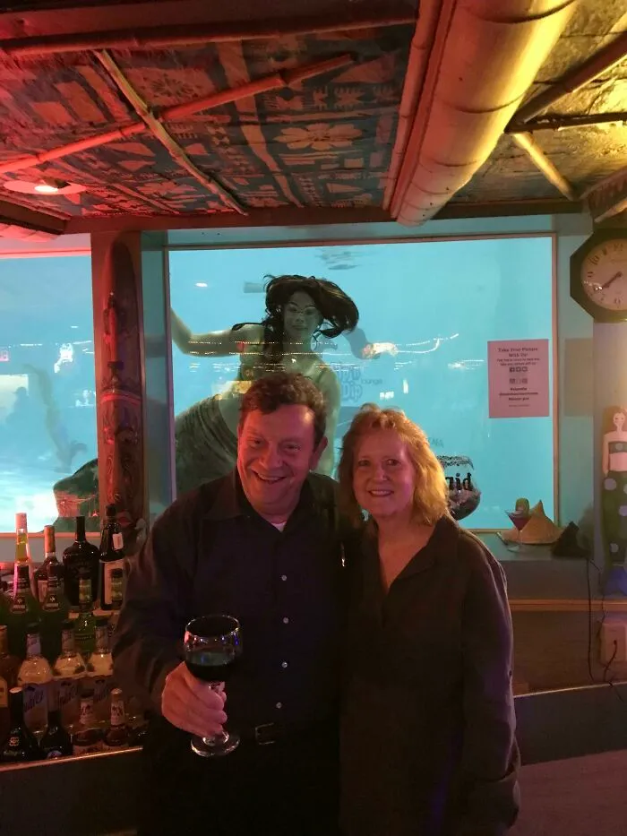 A couple smiling in a bar with a mermaid in the background tank