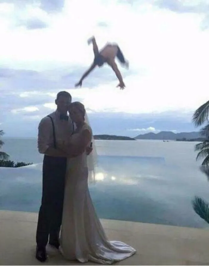 A man doing a backflip in the background of a romantic wedding couple photo by the water.
