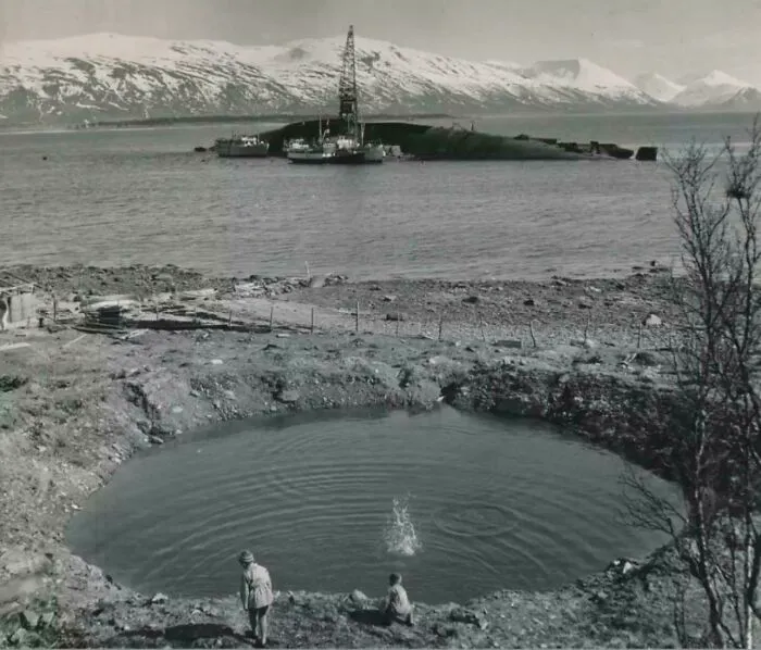 Two kids stand near a pond with a salvaged ship and mountains in the background