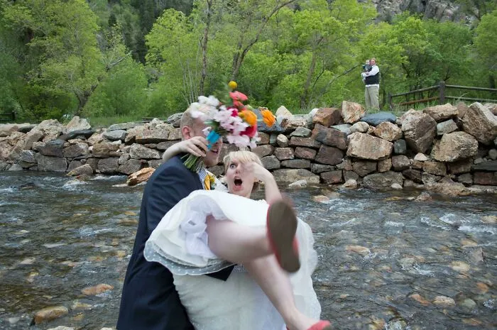 A groom carrying a bride who has a shocked expression as someone in the background watches them near a river.