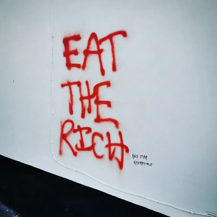 Red graffiti on a wall reading Eat the Rich with clever vandalism adding and stay hydrated.
