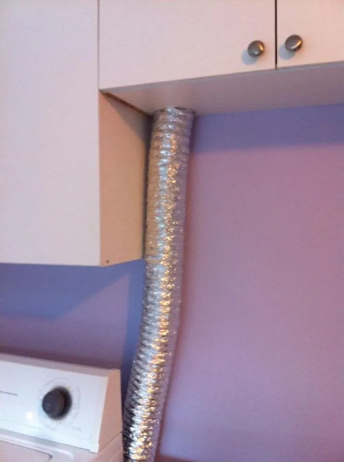 A flexible silver vent pipe awkwardly squeezed between white kitchen cabinets, illustrating a poor choice to renovate their homes.