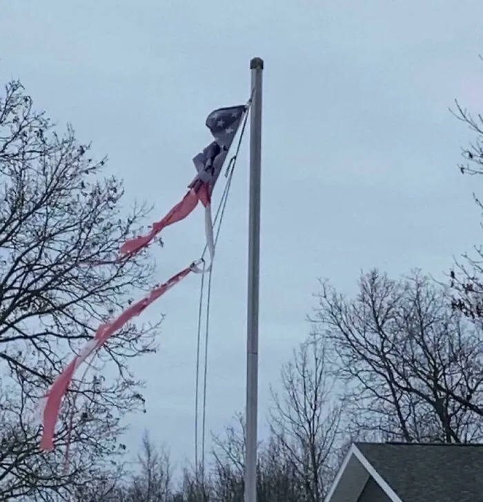 A tattered, shredded flag flying on a pole outdoors displaying neglect from infuriating neighbors