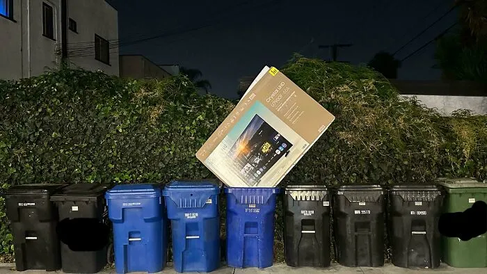 A row of trash cans outdoors with a large TV box leaning against them, annoying neighbors.