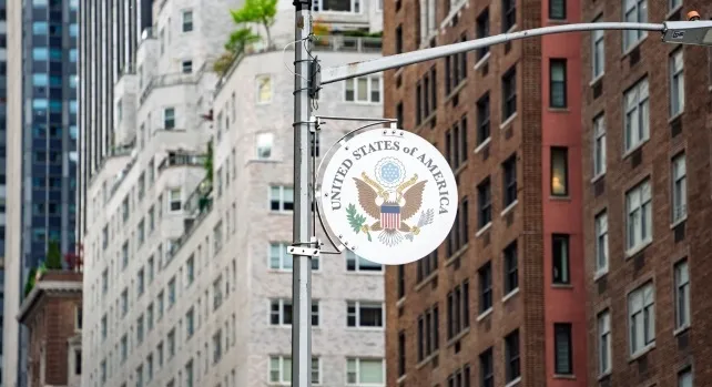 Round Great Seal of the United States hanging from a pole in front of city buildings representing US government.