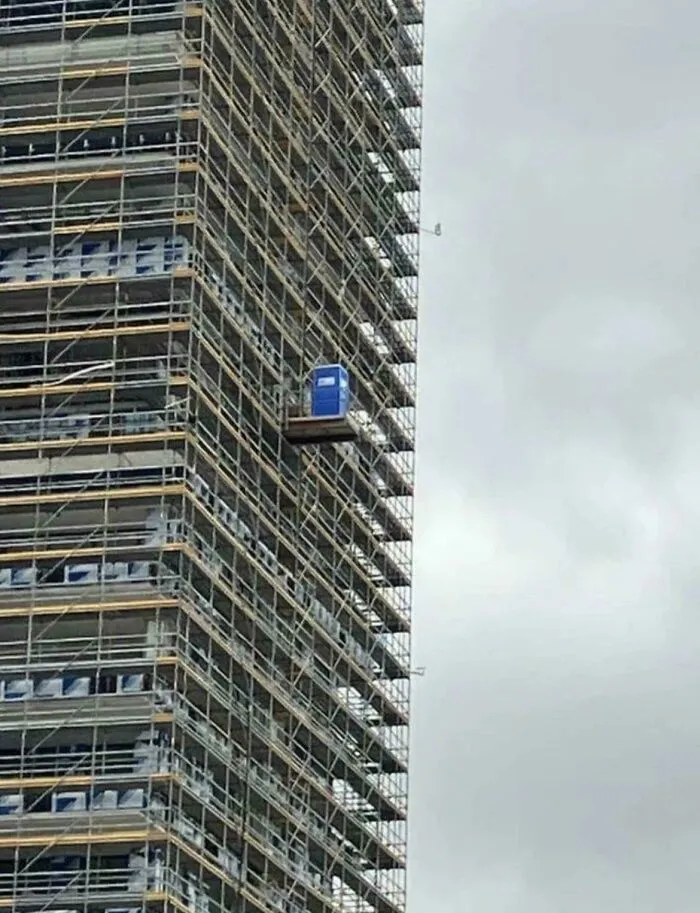 A porta potty suspended high up on a construction scaffolding platform in cursed images.