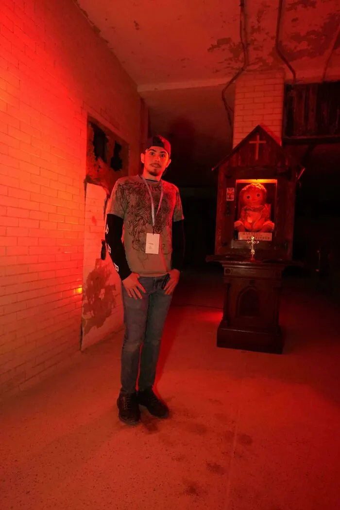Man standing near creepy Annabelle doll display in red illuminated room.