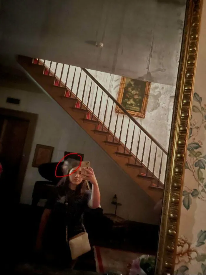 A girl taking a mirror selfie near a staircase with a shadowy figure behind her, featured in creepy photos.