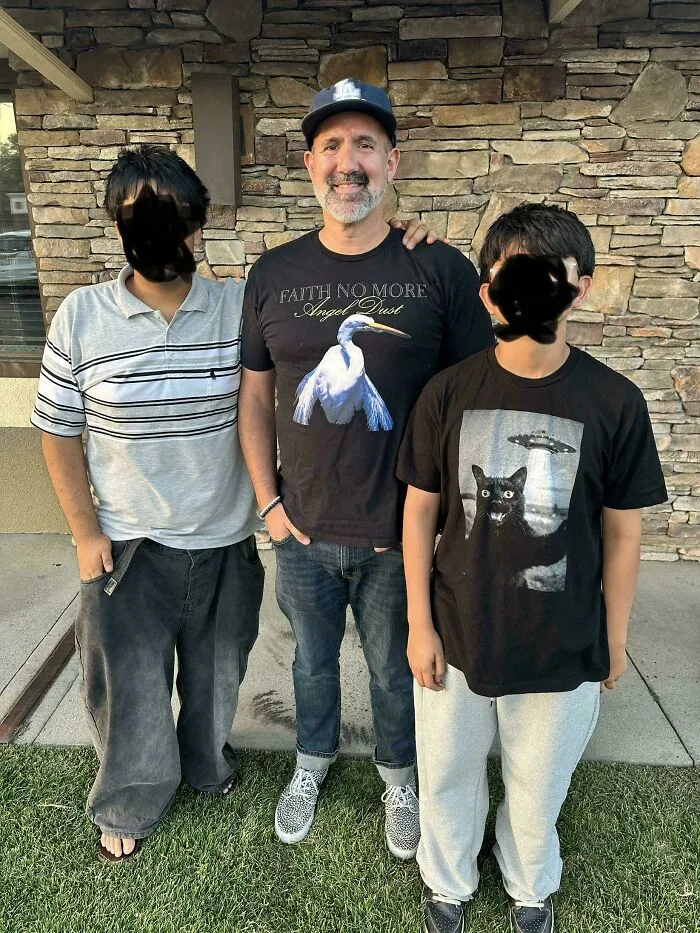 A father wearing a graphic shirt standing proudly outdoors between his two sons in a wholesome family photo.
