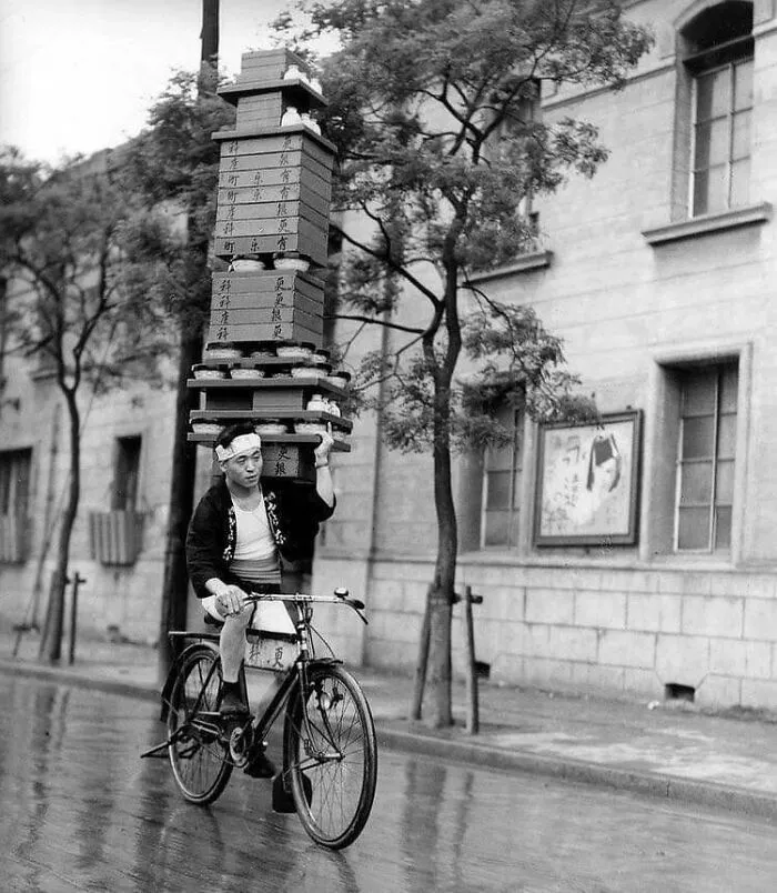 Black and white rare historical image of a man balancing tall stack on his head while riding a bicycle