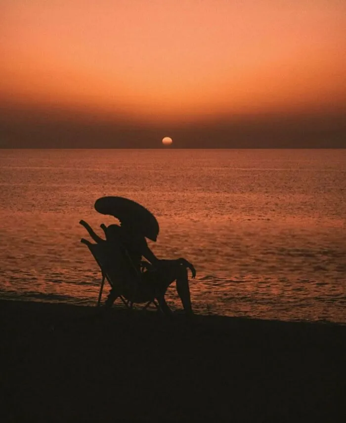 Silhouette of a Xenomorph creature sitting on a beach chair during sunset in weird photos.