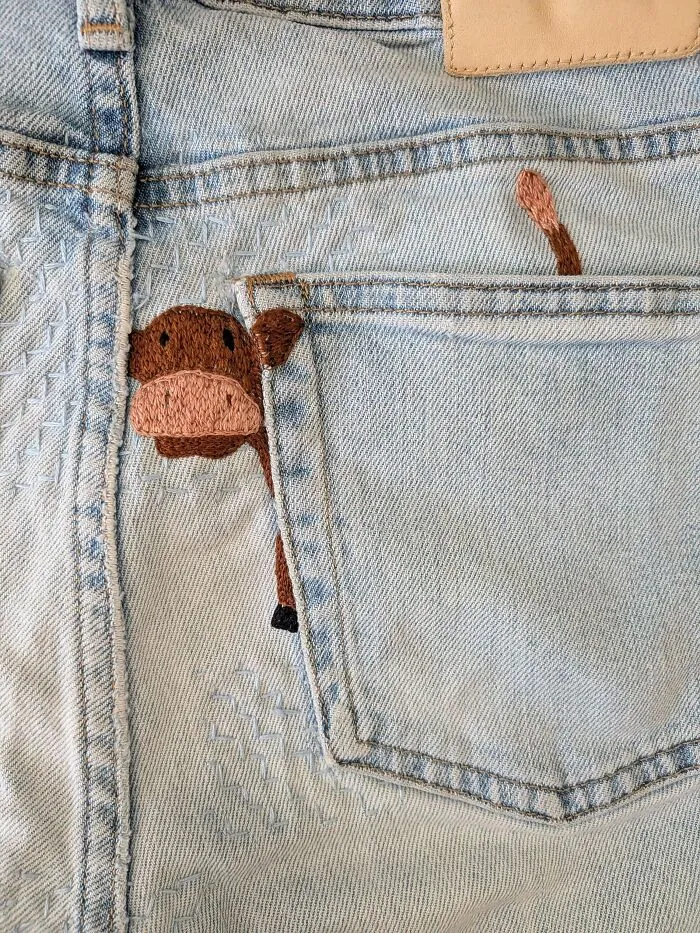 A cute brown cow embroidered peeking out of a denim jeans pocket using creativity.