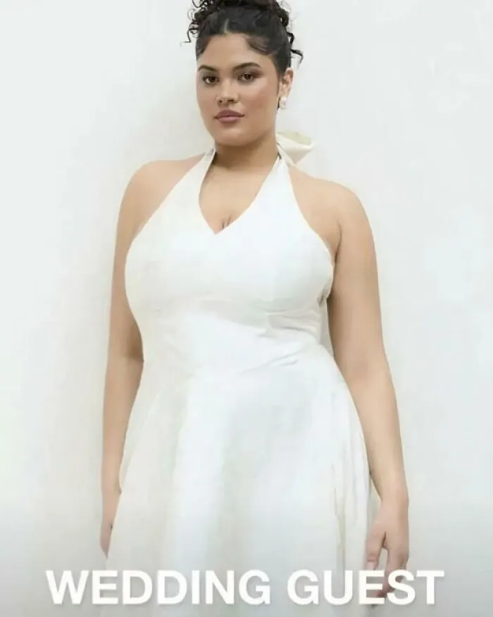 Woman wearing white dress labeled wedding guest posing against plain background