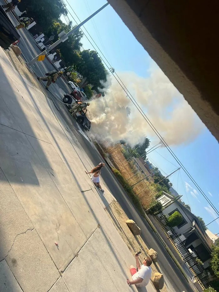 Influencers posing for content in public spaces while firefighters tackle a burning car nearby.