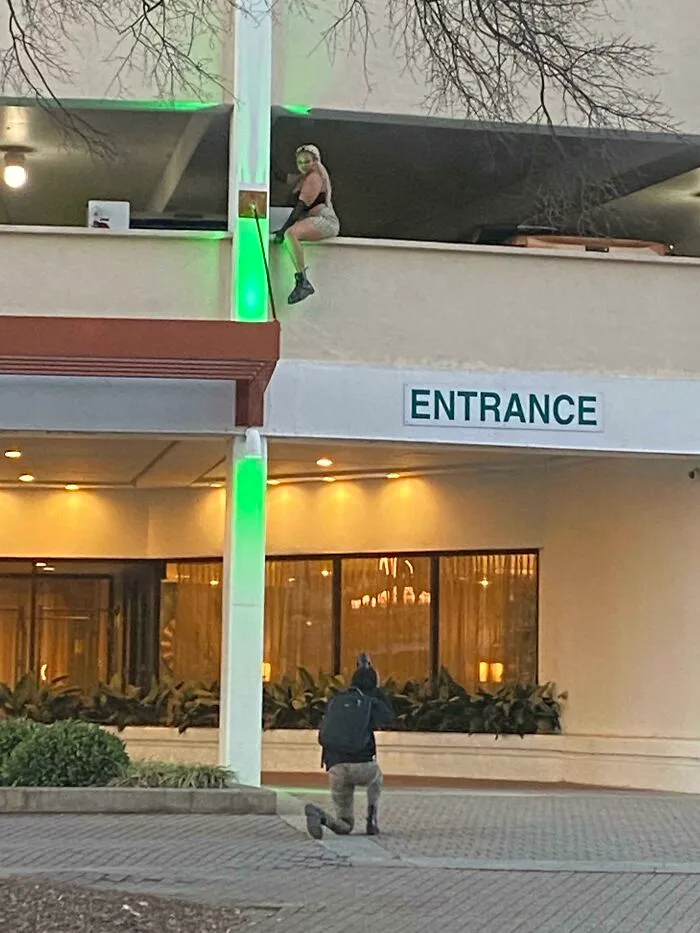 A photographer kneels on a public sidewalk taking pictures of an influencer sitting dangerously on a parking ledge.