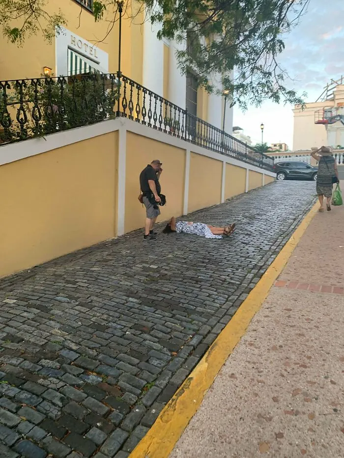 An influencer lies on a public cobblestone street while a photographer stands over her taking photos.