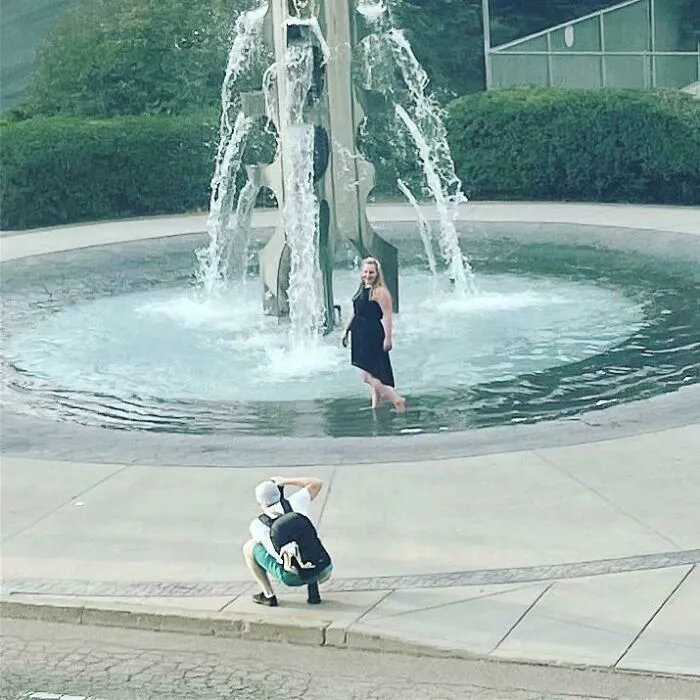 A photographer crouches in public spaces to capture an influencer posing inside a large outdoor water fountain.