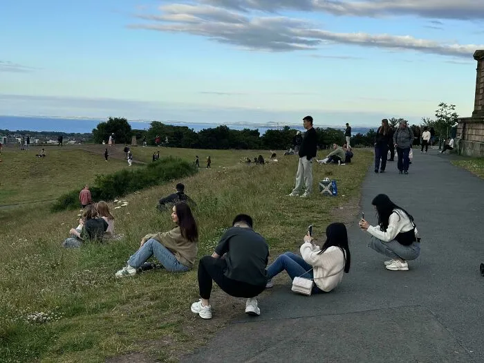 Group of influencers in public spaces taking photos with smartphones on a grassy hill overlooking the sea.