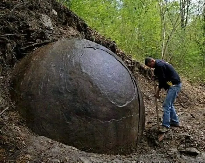 A man standing next to a massive, spherical stone embedded in the ground, showcasing surprising facts about the past.