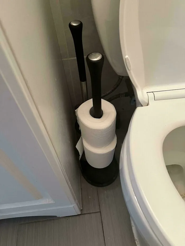 Spare toilet paper rolls stacked onto the handle of a toilet plunger next to the bowl.