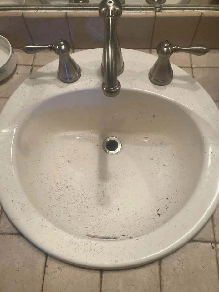 A white bathroom sink covered in tiny hair trimmings after a partner shaved.